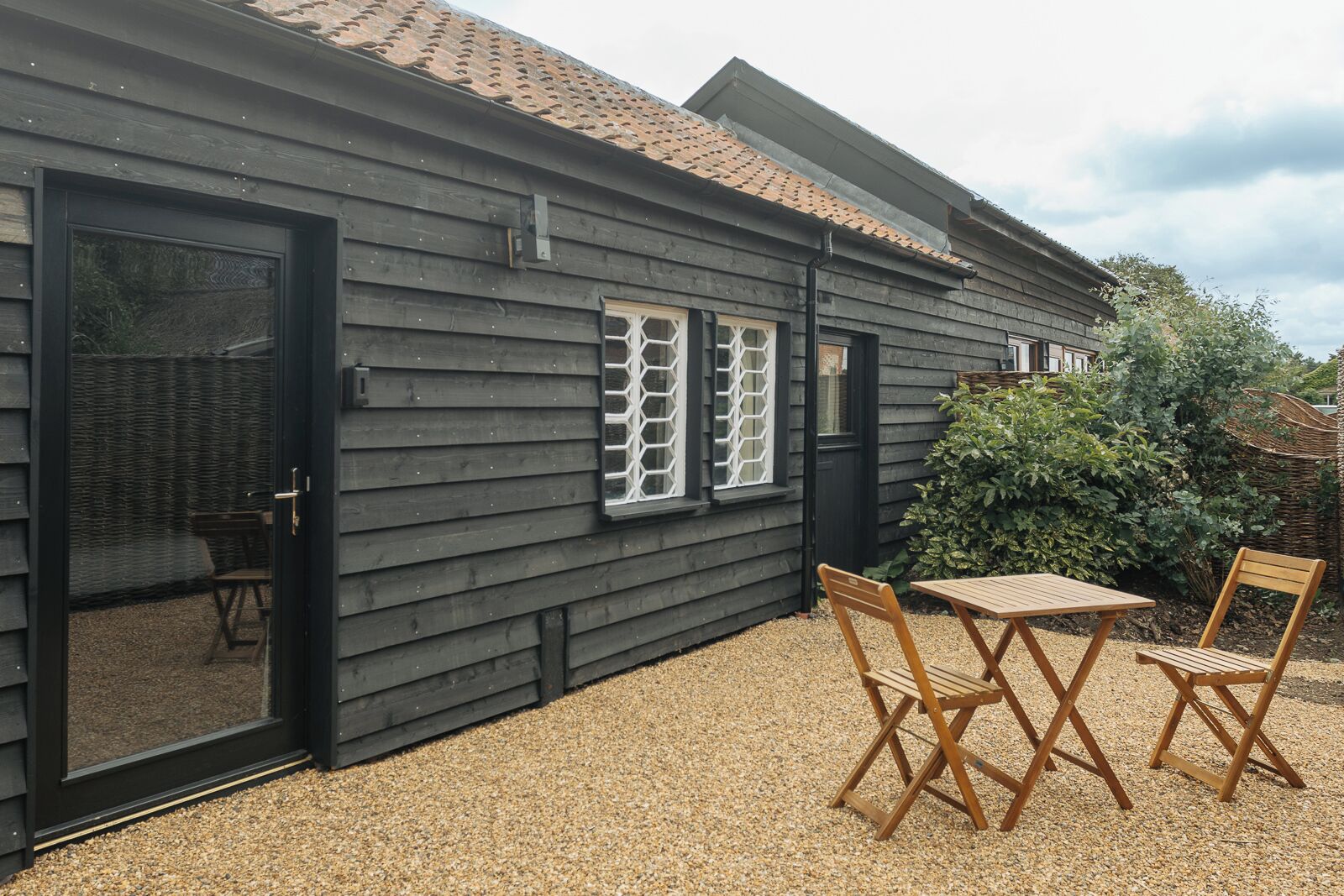 Il s’agit d’un bâtiment en bois à l’extérieur noir, doté d’une porte coulissante en verre et d’un petit espace patio avec deux chaises et une table en bois.