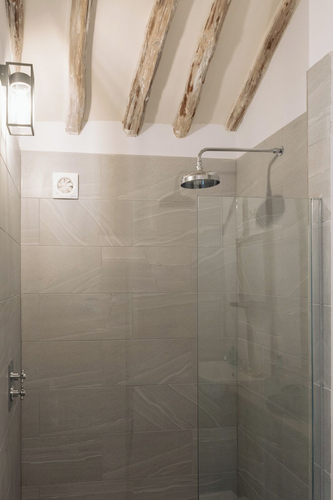 Une douche fermée par des parois en verre, dotée d’un pommeau de douche à main, d’un luminaire fixé au mur et d’une grille d’aération visible.