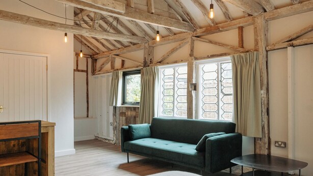 A rustic room with exposed wooden beams, a green sofa, a small table, and a window with curtains.
