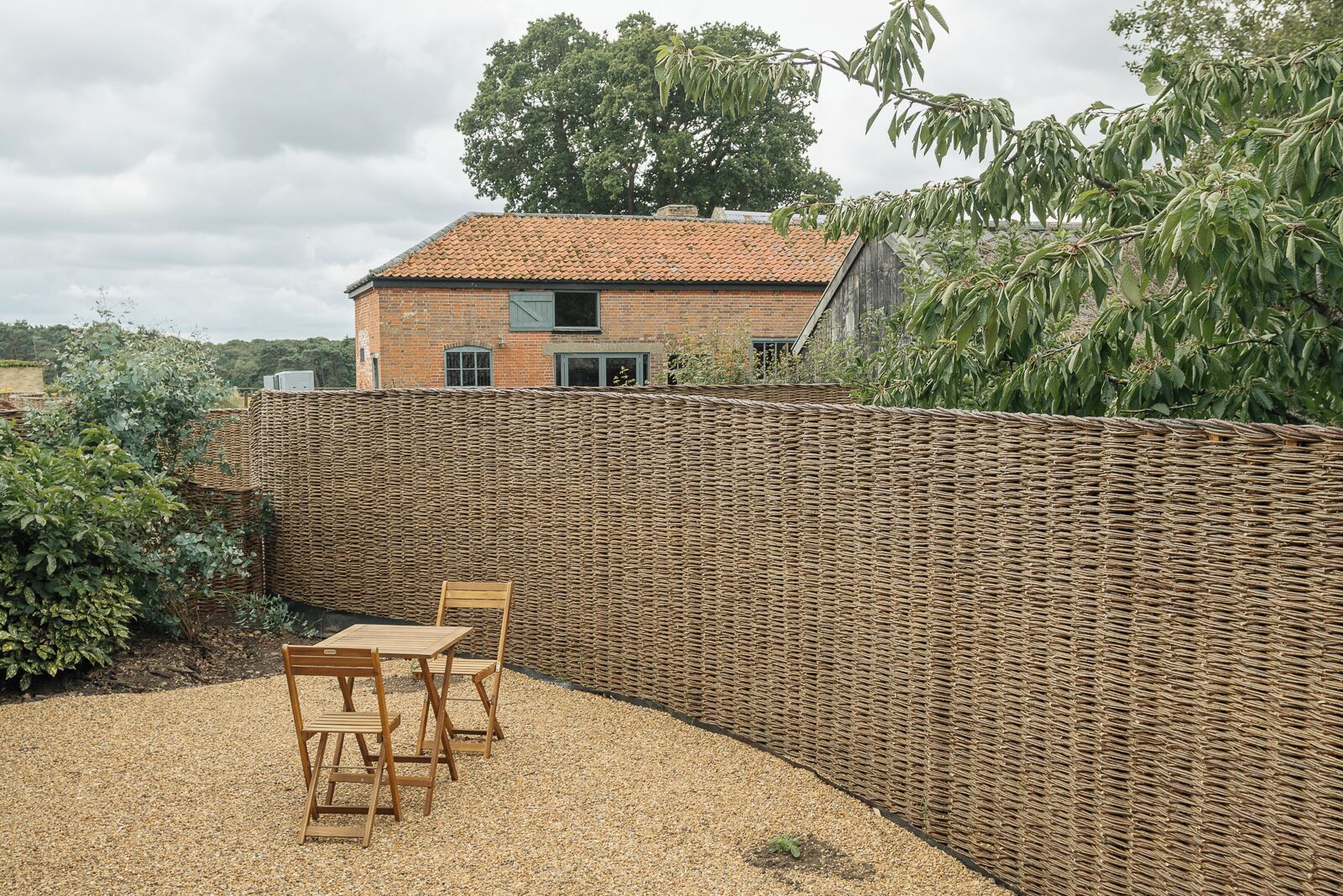 A patio with a wicker wall, a brick house in the background, and a gravel ground.