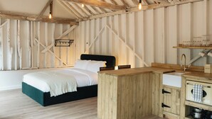 A rustic bedroom with exposed wooden beams, a bed with a black headboard, a wooden desk, and a vintage-style kitchen area.