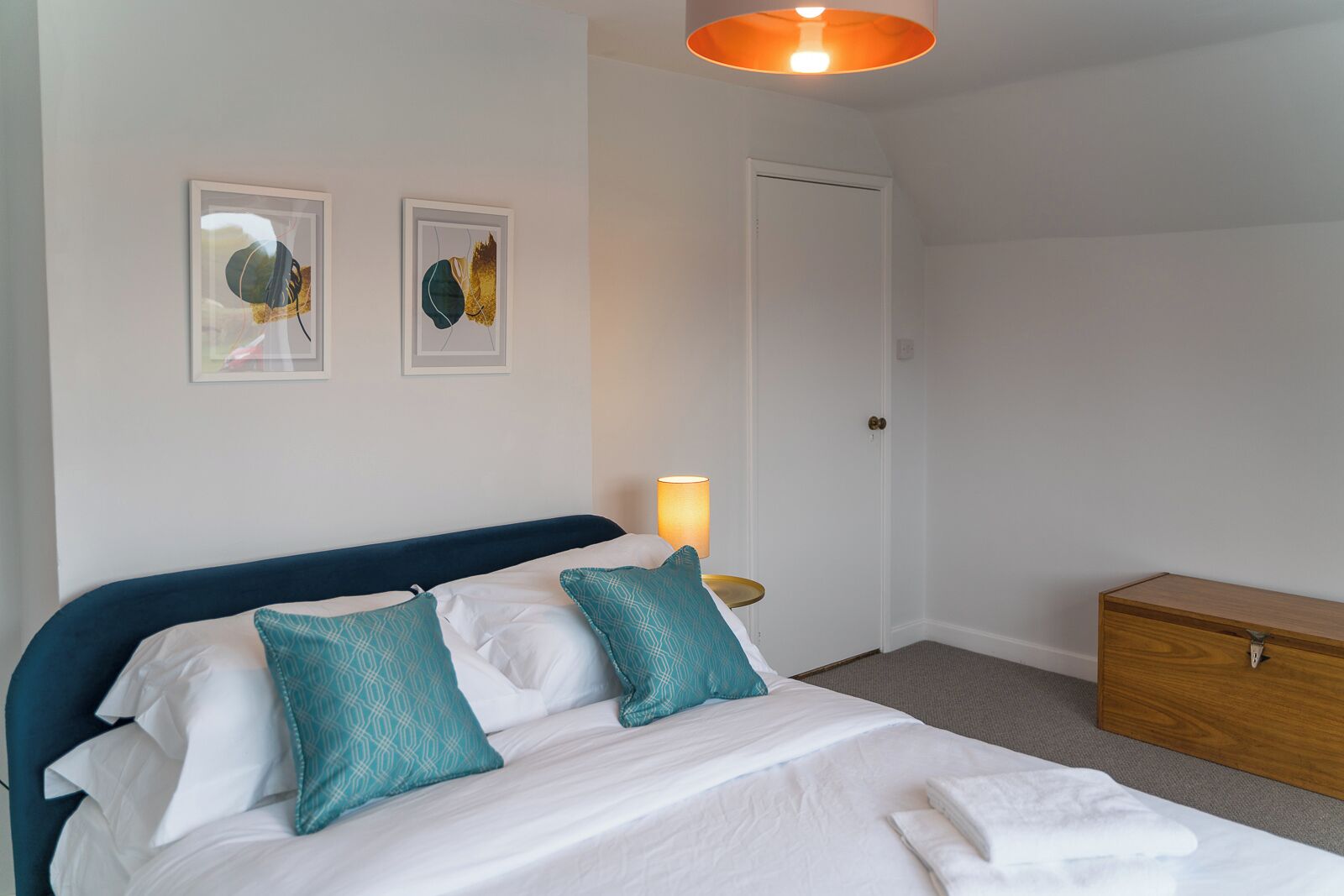 Un lit bien fait, avec du linge de lit blanc et des coussins turquoise, une lampe de chevet et une commode en bois, dans une pièce simple et épurée.