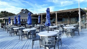 Ein Außenbereich mit Essbereich, der runde Tische und Stühle sowie blaue Sonnenschirme und eine Holzterrasse umfasst.