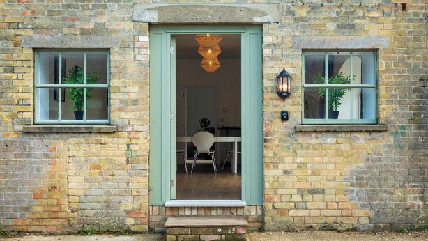 A brick building with a green door, two windows with plants, and a hanging light fixture. - Long Range 1, Henham Park Nr Southwold (Beccles)