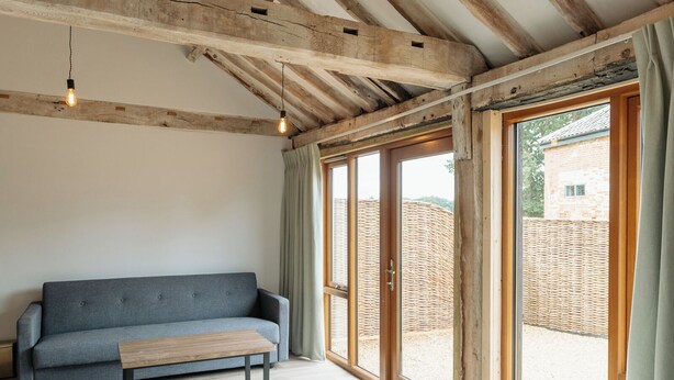 A modern living room with a grey sofa, a wooden bench, and large windows offering a view of a brick wall and greenery outside. - 4 Henham Barns, Henham Park nr Southwold, NR34 8AQ, Carefully restored Character filled barn (Beccles)