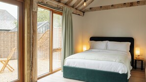 A bedroom with a large bed, wooden beams, a wicker chair, and a view of a building outside. - 4 Henham Barns, Henham Park nr Southwold, NR34 8AQ, Carefully restored Character filled barn (Beccles)