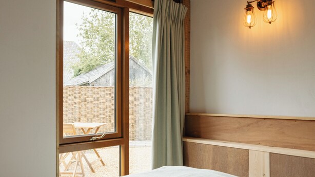 Une chambre à coucher avec une tête de lit en bois, une fenêtre donnant sur une table en osier à l’extérieur, et un luminaire fixé au mur.