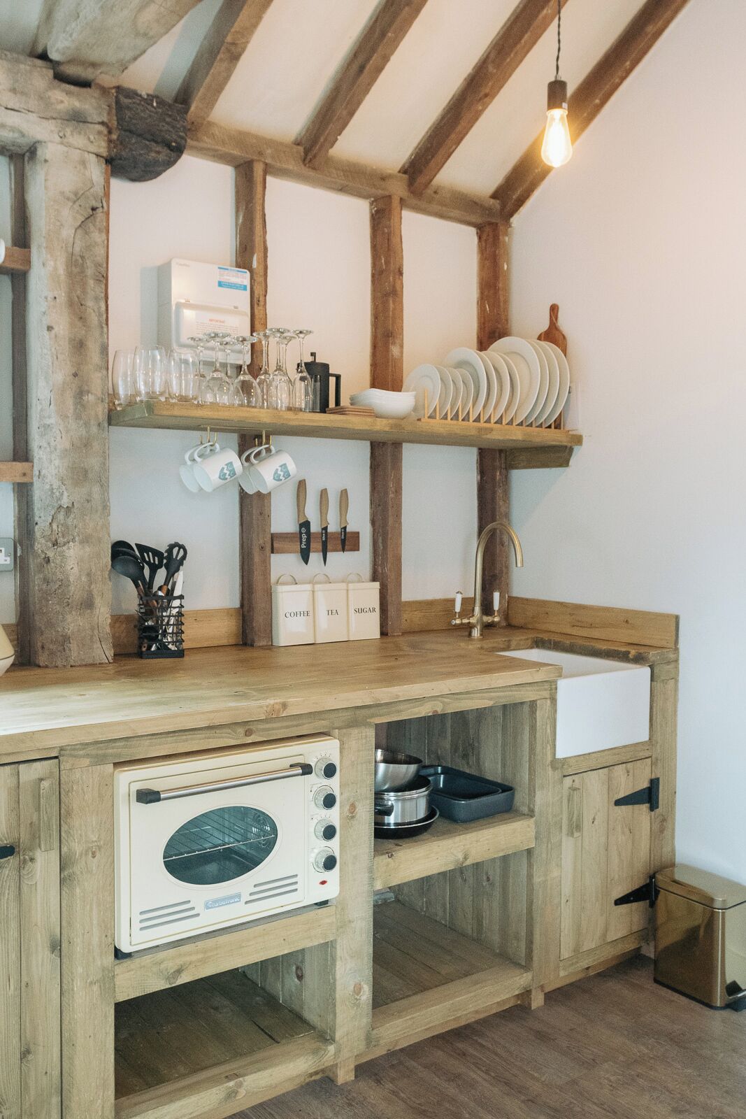 A rustic kitchen with wooden beams, a built-in oven, and shelves holding dishes and utensils.