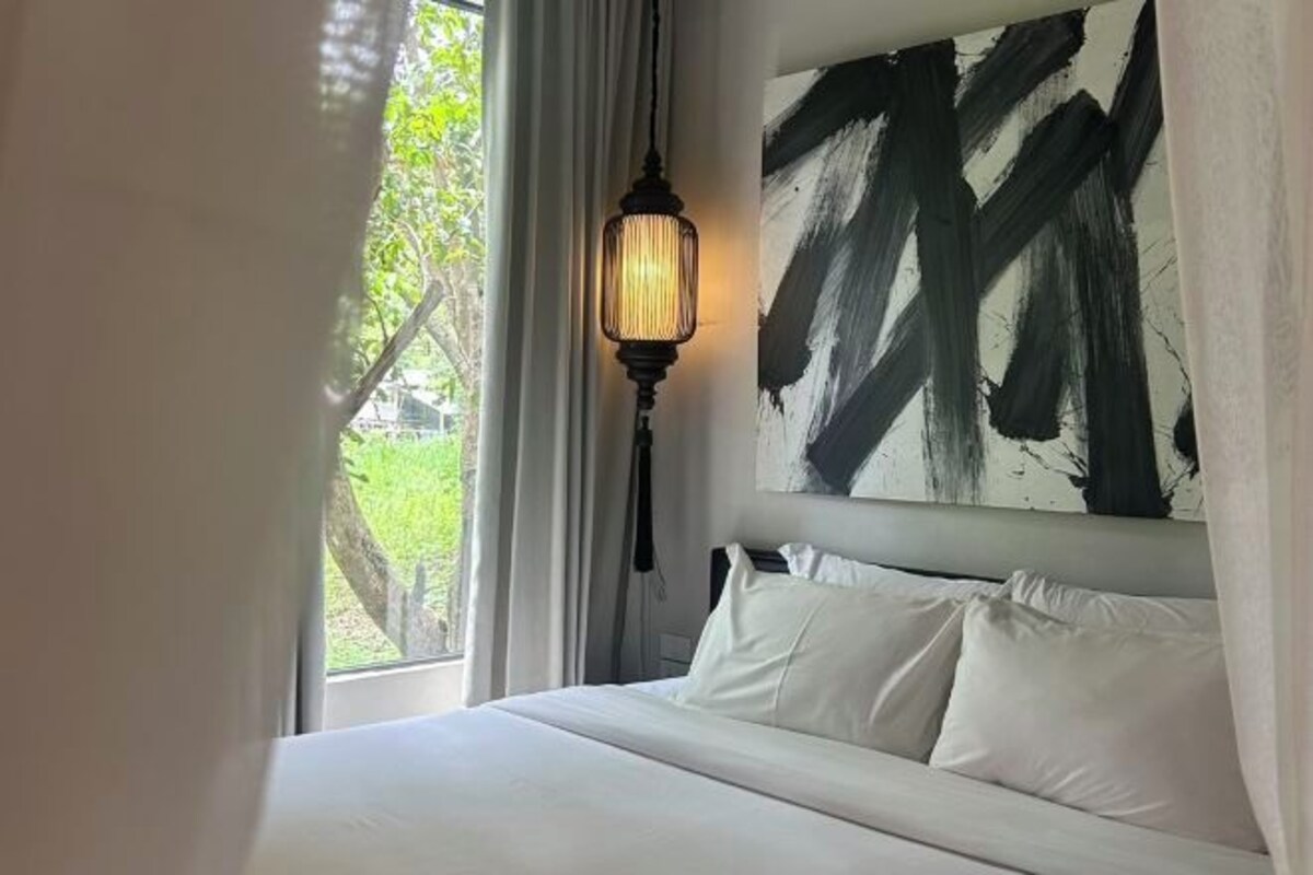 Quarto com cama, roupa de cama branca, pintura abstrata preta, luminária pendente e vista para o verde lá fora.