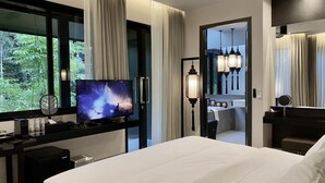 A modern hotel room with a large bed, a TV on a stand, a desk with a computer, and a bathroom visible through an open door.