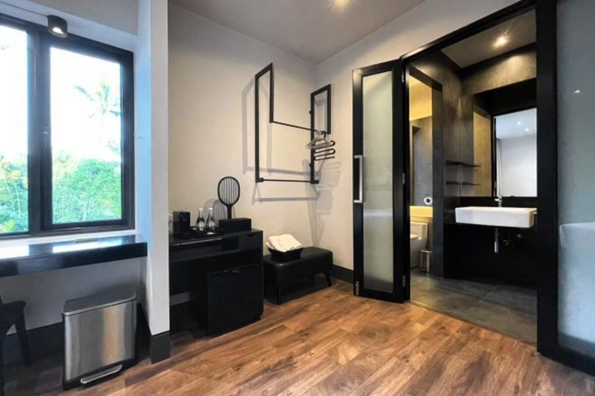 Quarto moderno com piso de madeira, escrivaninha preta, lixeira pequena, cadeira preta, espelho e banheiro com pia e vaso sanitário.