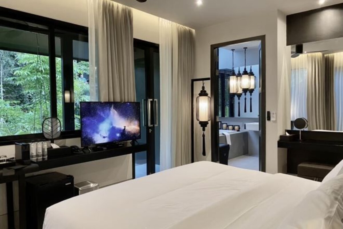 Quarto de hotel moderno com uma cama grande, uma televisão em uma escrivaninha, um banheiro com espelho e iluminação, e vista para o verde lá fora.