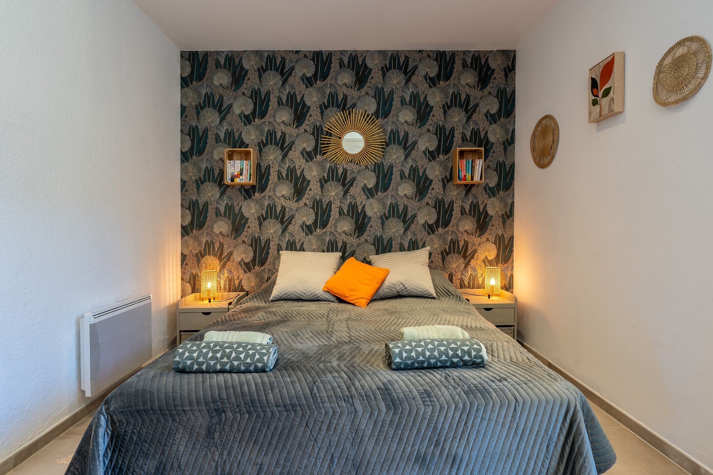 A bedroom with a patterned wallpaper, a bed with a grey quilt, two bedside tables with lamps, and a wall-mounted mirror.