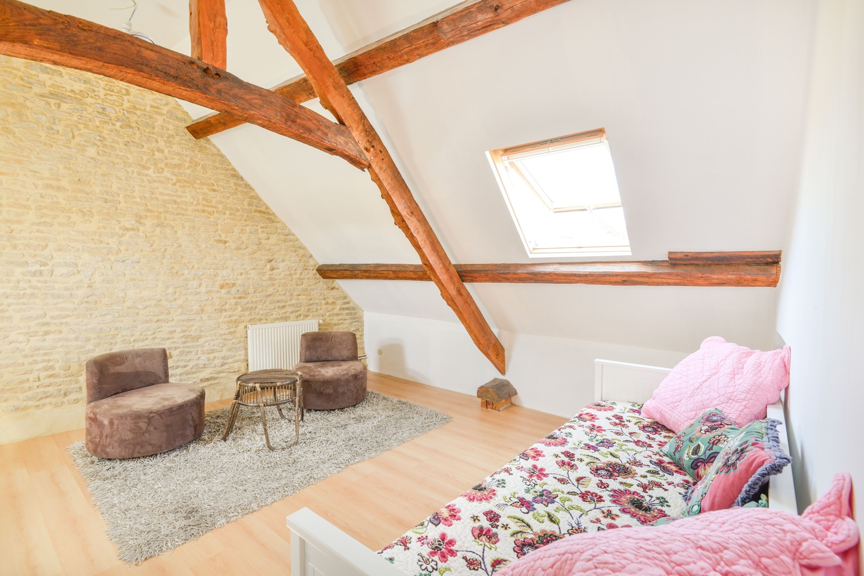Une chambre mansardée chaleureuse avec des poutres apparentes, un mur en pierre, une petite table avec des chaises, un lit avec une literie à motifs floraux et un puits de lumière.