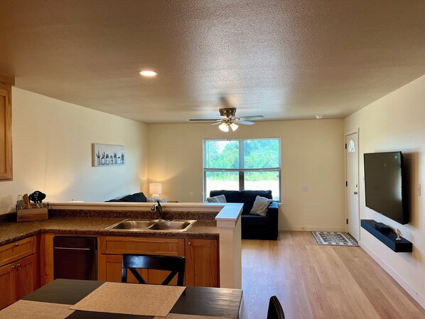 A modern kitchen with granite countertops, a dining area with a table and chairs, a living room with a sofa, a large window, and a ceiling fan.