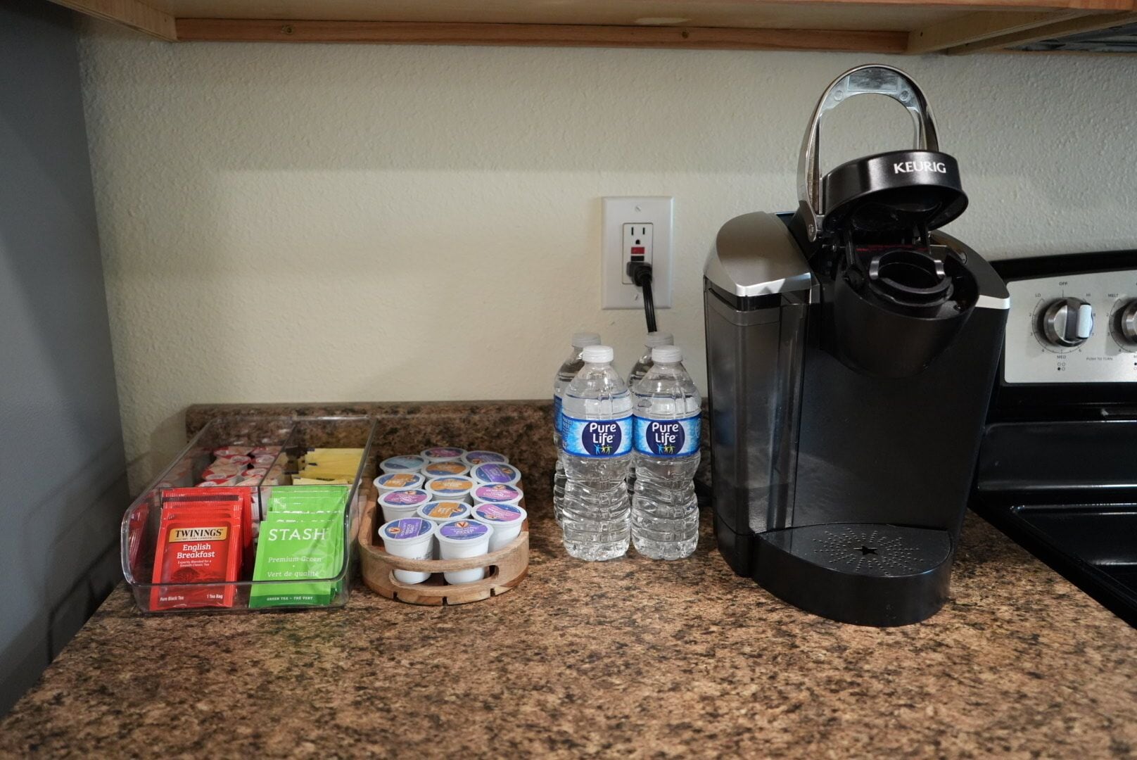 Een keukenblad met een Keurig koffiezetapparaat, Twinings English Breakfast thee, groene thee van Stash, Pure Life waterflessen en een houten mand met eieren.