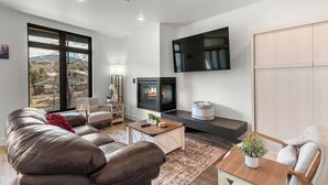 A modern living room with a leather sofa, a coffee table, a fireplace, and a large TV mounted on the wall. - Serenity in Silverthorne: Luxury Condo Retreat with Mountain Views (Silverthorne)