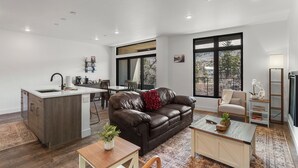 A modern living space with a kitchen, a brown leather sofa, a dining area, and large windows offering a view of the outdoors. - Serenity in Silverthorne: Luxury Condo Retreat with Mountain Views (Silverthorne)