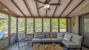 A covered outdoor seating area with a wooden ceiling, a ceiling fan, a cushioned sofa with pillows, a wooden table, and a view of trees through large windows. - Rustic Fox by Innsbrook Vacations! (Innsbrook)