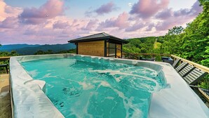 Een jacuzzi met een houten terras en lounge stoelen, omgeven door weelderige begroeiing en bergen op de achtergrond.
