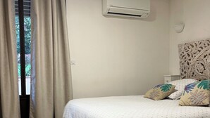 Un dormitorio con cama, cortinas color beige, aire acondicionado, piso de madera y una mesita de noche con lámpara.