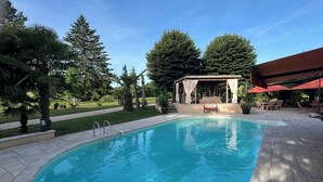 Una piscina con solárium de piedra, zona de estar cubierta y abundante vegetación.