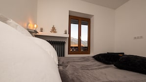 A bedroom with a bed, a nightstand with a lamp, a window with a view of mountains, and a fireplace. - Chalet les Myrtilles (Venthon)