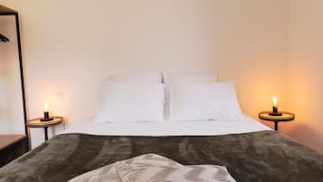 Un lit bien fait, avec du linge de lit blanc et une couverture marron foncé, flanqué de deux tables de chevet sur lesquelles sont posées des bougies.