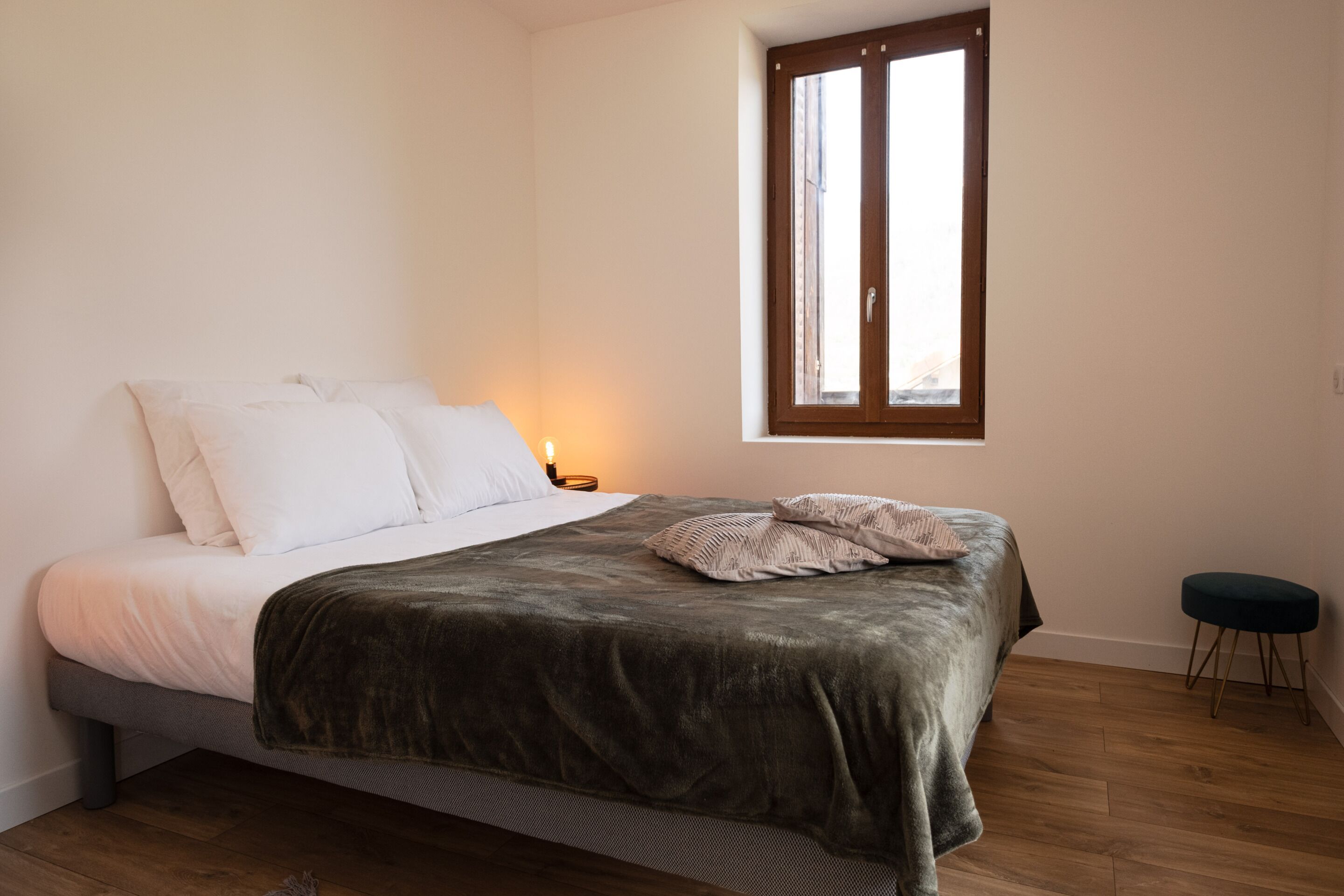 A bedroom with a bed, a window, a bedside table, and a small stool.