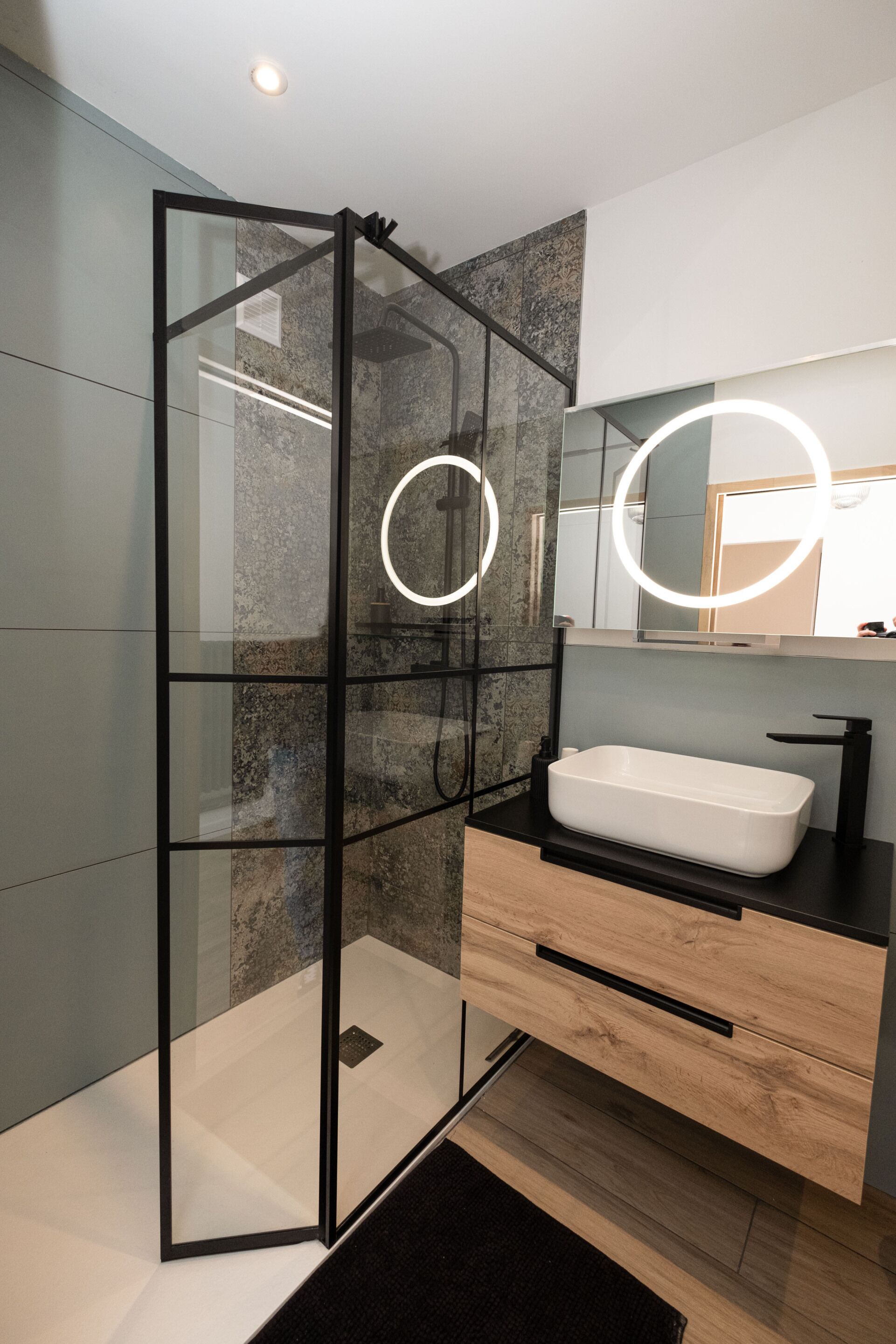 Un baño moderno con ducha envuelta en vidrio, lavamanos blanco y espejo con luces circulares.
