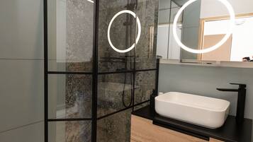 Un baño moderno con ducha envuelta en vidrio, lavamanos blanco y espejo con luces circulares.