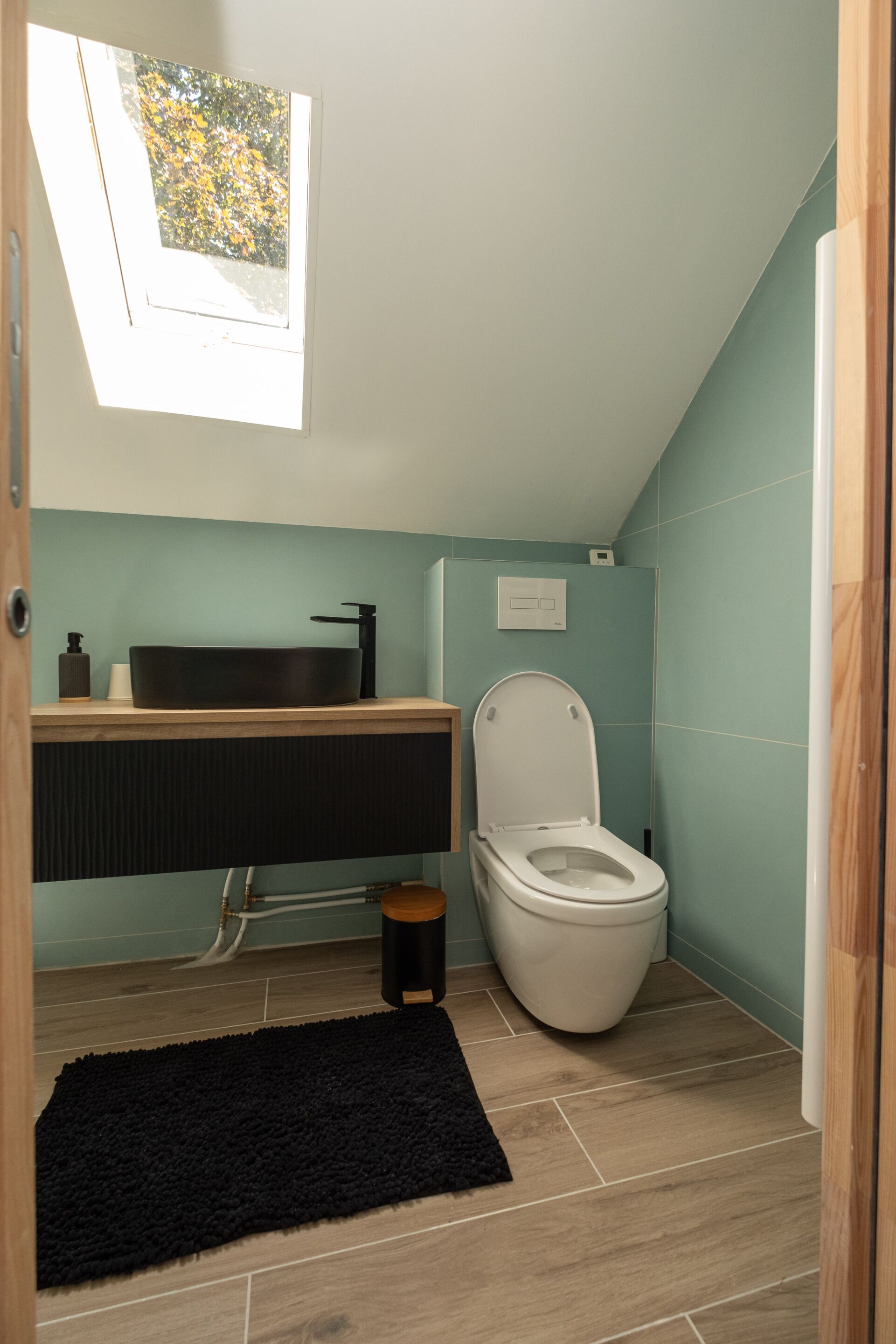 Een compacte badkamer met een dakraam, een zwart vloerkleed en een moderne wastafel.