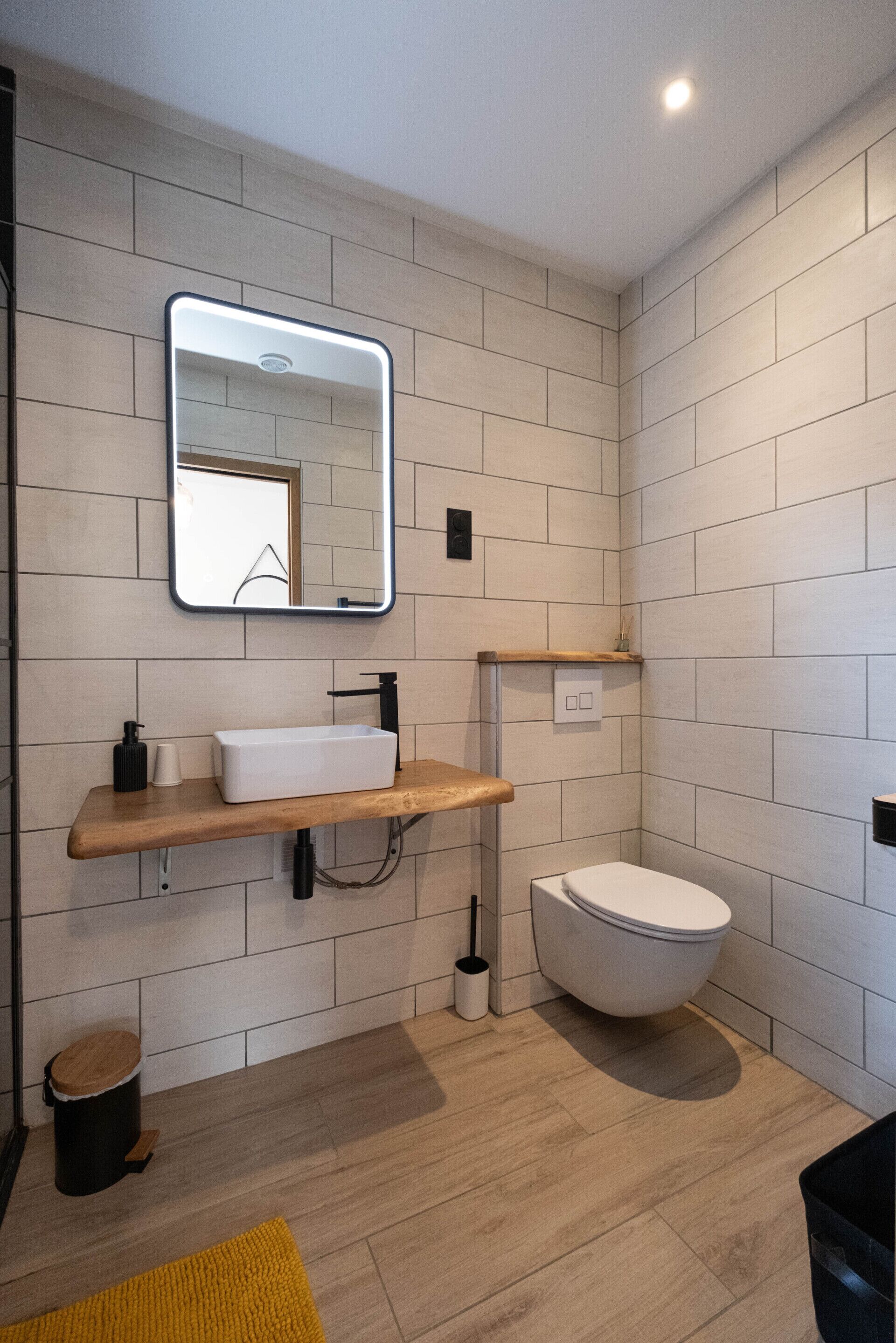 Un baño moderno con pared de azulejos blancos, tocador de madera, lavamanos blanco, grifo negro, inodoro blanco y espejo con luz.