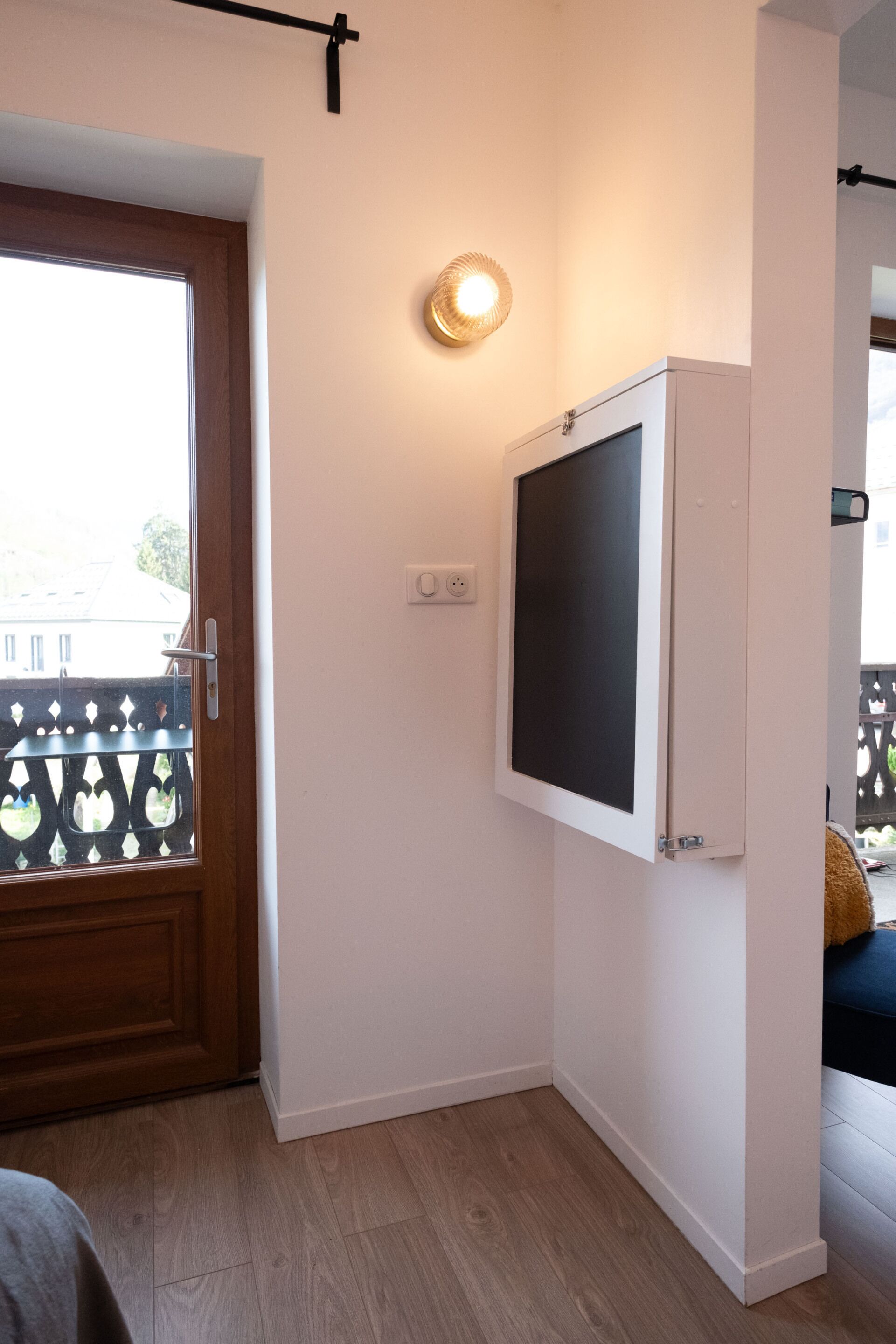 Una habitación con puerta de madera, televisor blanco montado en la pared, lámpara y vistas a través de una ventana con celosía decorativa.