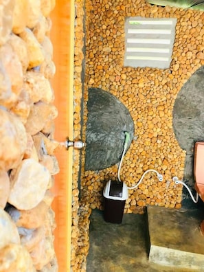 A room with a stone wall, a metal door, a black trash bin, and a power outlet with a charging cable. - Atha Safari Resort & Riverside Camping (Udawalawa)