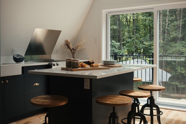 A modern kitchen with a large island, bar stools, and a view of a forest through a large window.