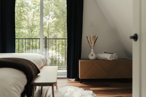 A bedroom with a bed, bench, bedside table, and a view of greenery through a window. - Kartalla - Aframe chalet lake access near Tremblant (Amherst)