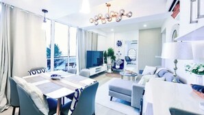 A modern living room with a dining area, large windows, a flat-screen TV, and a chandelier. - Lazzlla 1BR beachfront oceanview apartment (Hulhumale city Kaafu)