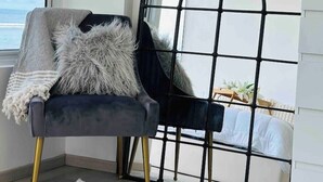 A room with a view of the sea, a black metal-framed window, a grey upholstered chair with a fluffy pillow, and a white dresser. - Lazzlla 1BR beachfront oceanview apartment (Hulhumale city Kaafu)