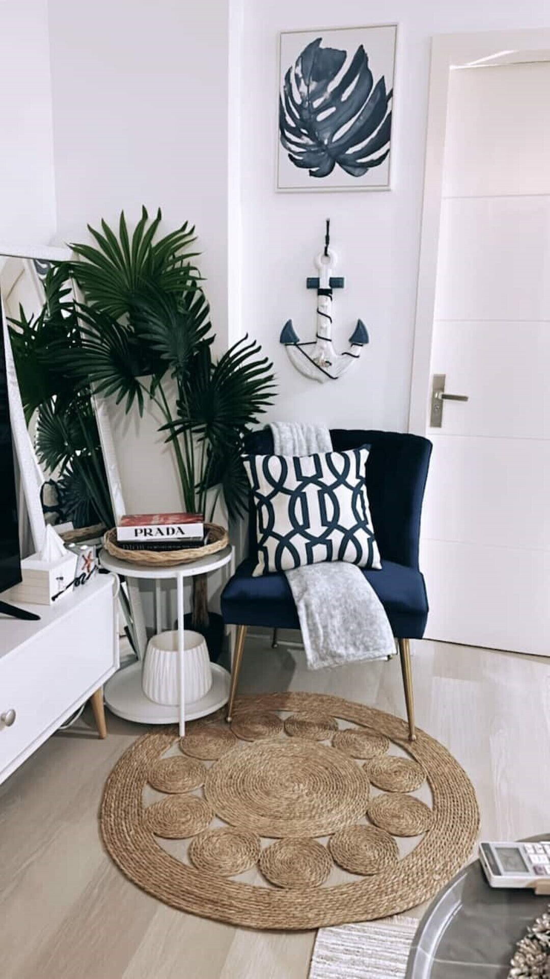 A modern living room with a dark blue armchair, a round woven rug, a white side table with a book, and a large plant.