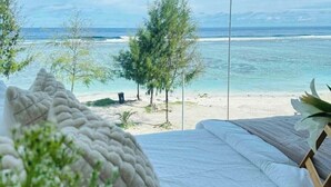 A bedroom with a bed facing a beach, a clear view of the ocean, and a few trees in the distance. - Lazzlla 1BR beachfront oceanview apartment (Hulhumale city Kaafu)