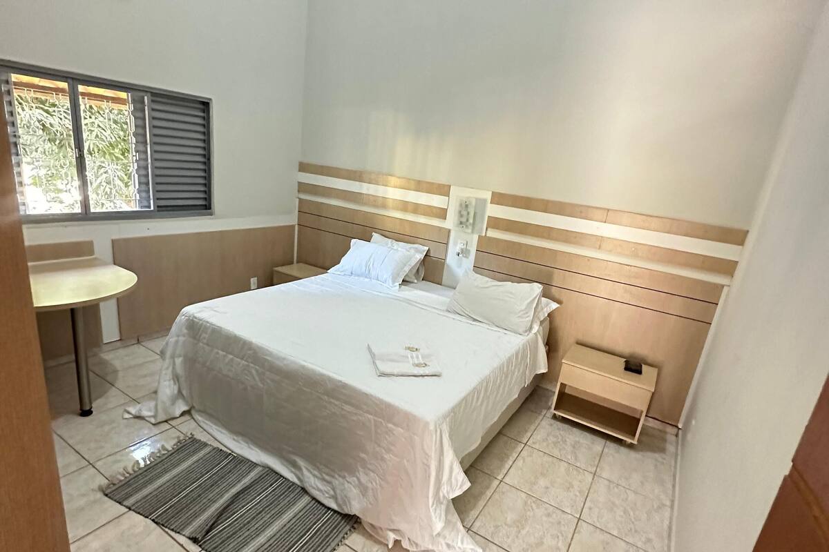 Habitación de hotel con cama, mesita de noche y ventana con persianas.