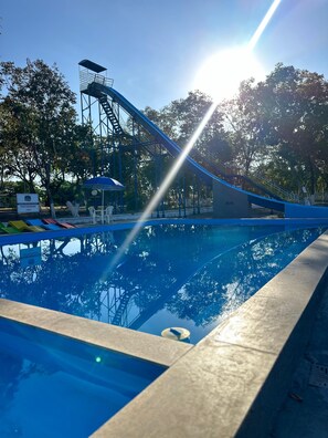 Une piscine avec un toboggan bleu, entourée d’arbres et sous un ciel dégagé.