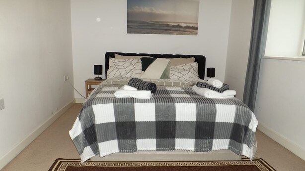 A neatly made bed with a checkered bedspread, two pillows, and a folded towel. There is a small bedside table with a lamp, a wall-mounted picture, and a window with curtains. - Charming 1-bed Apartment in Barking (Barking)