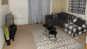 A modern living room with a sectional sofa, a black coffee table, a patterned rug, a TV on a stand, and a decorative vase with flowers. - Charming 1-bed Apartment in Barking (Barking)