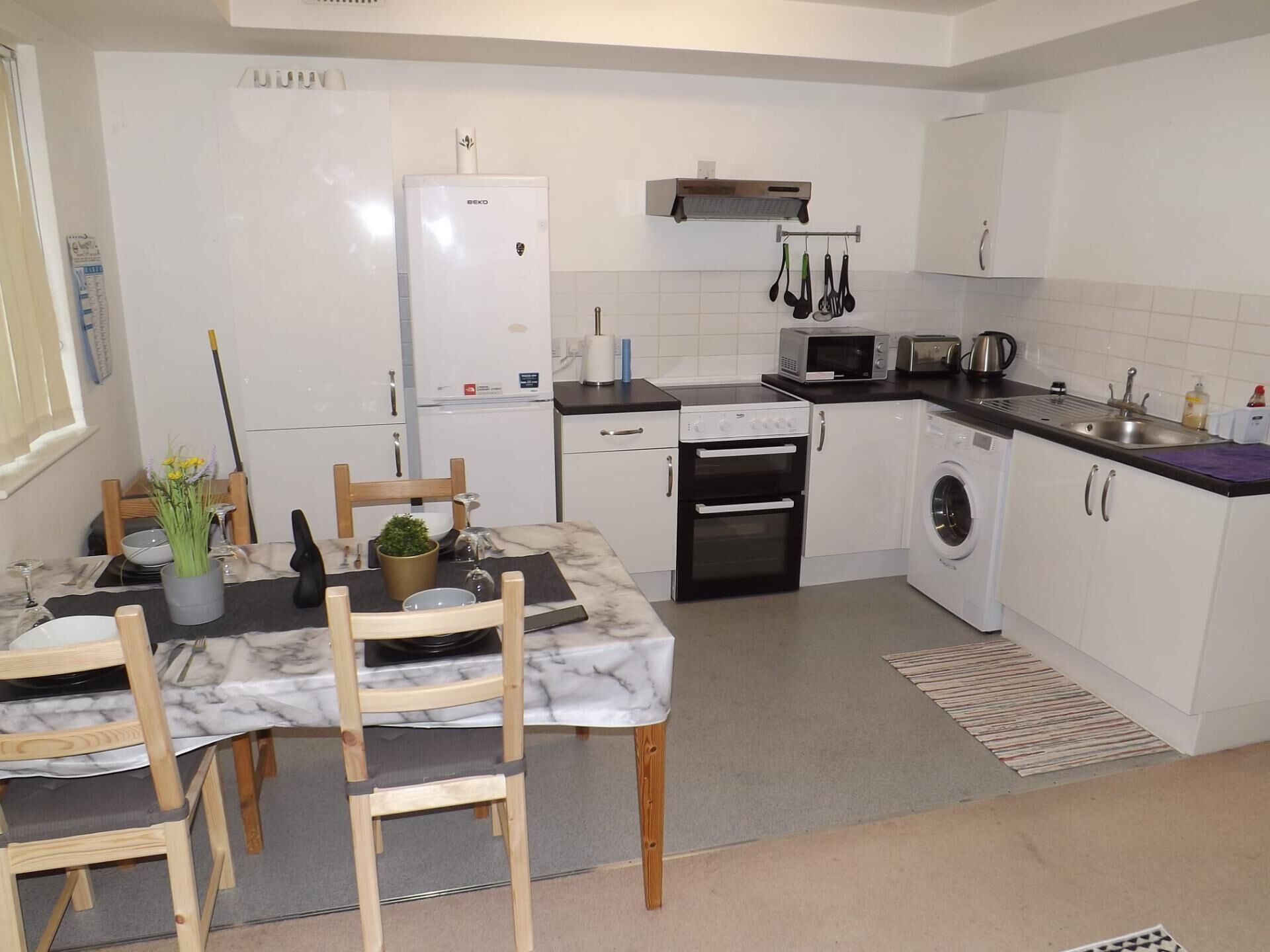 A modern kitchen with white cabinets, a built-in oven, microwave, and a washing machine. There is a dining table with chairs and a vase of flowers.
