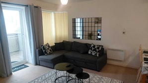 A modern living room with a grey sectional sofa, two black coffee tables, a patterned rug, a large mirror, and a sliding glass door leading to a balcony. - Charming 1-bed Apartment in Barking (Barking)
