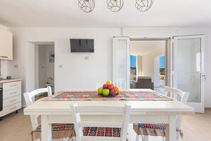 A dining area with a white table, chairs, and a fruit bowl. There is a TV mounted on the wall and a door leading to an outdoor area. - 3863 Dimora Le Frasche - Trilo 1 by Barbarhouse (Porto Cesareo)
