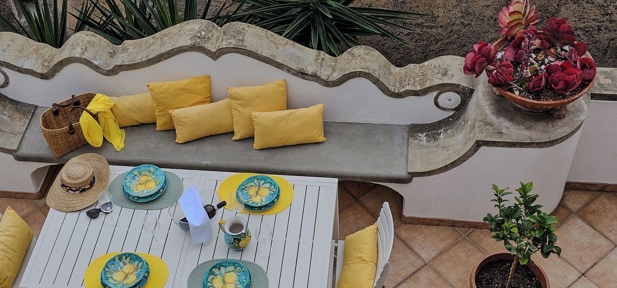 A patio with a white table set with yellow and blue plates, a wicker basket, and a potted plant.