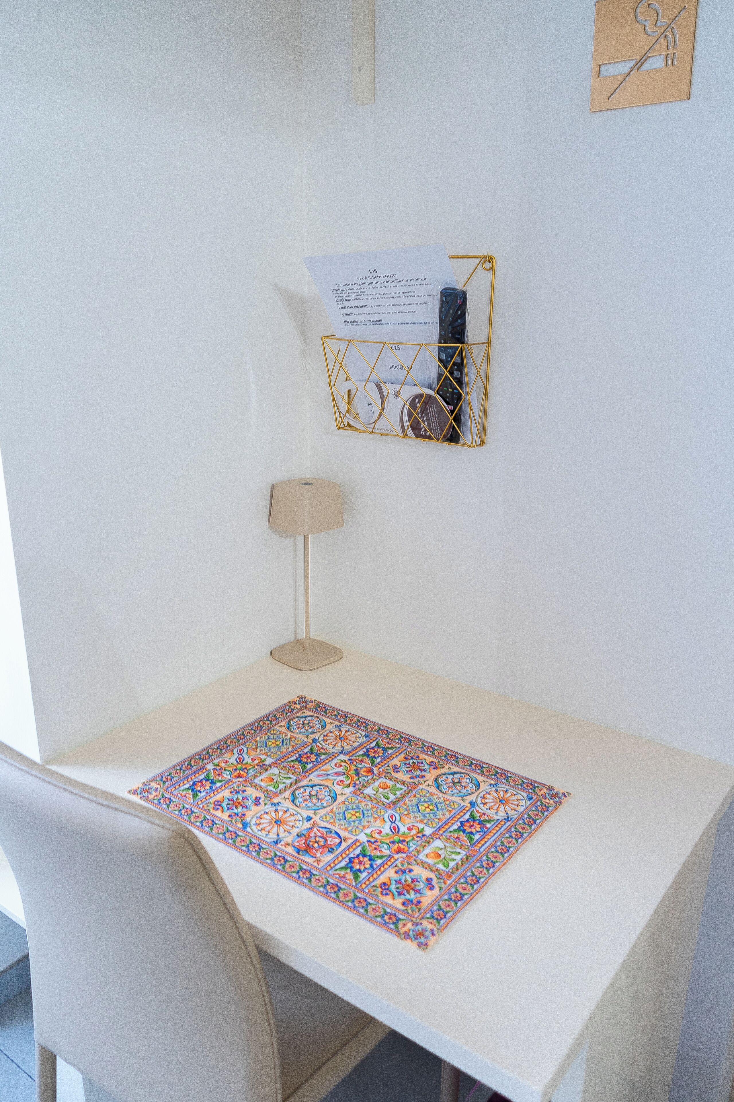 Un escritorio blanco con un mantel decorado, una lámpara y un organizador de pared con papeles y un teléfono.
